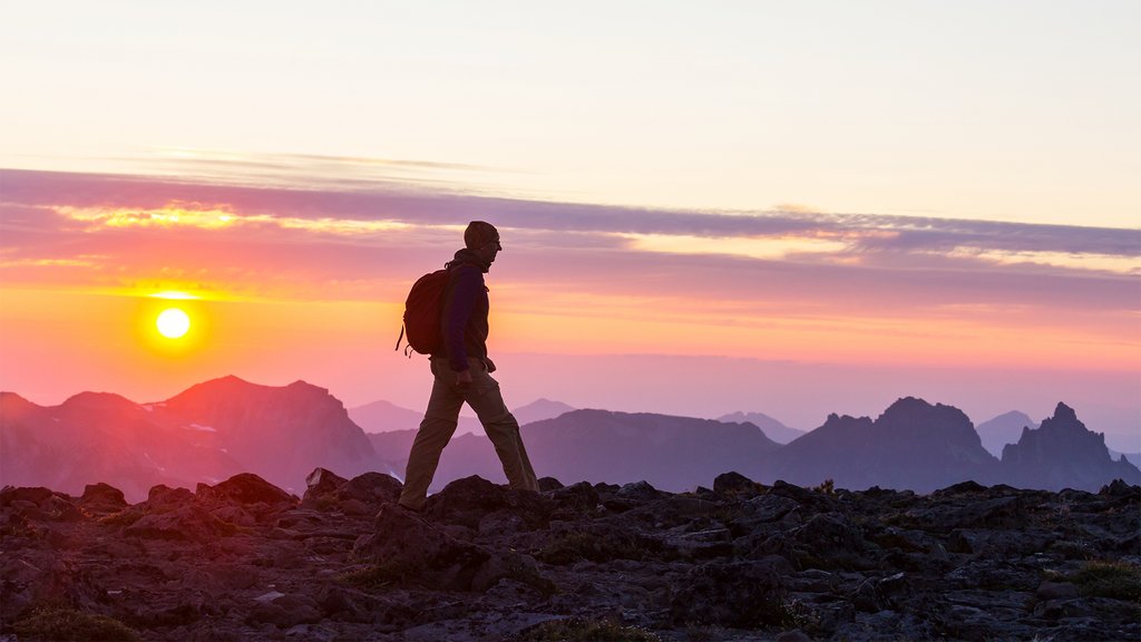Person wandert bei Sonnenuntergang auf einer felsigen Berglandschaft mit Berggipfeln im Hintergrund.